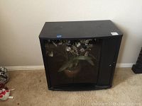 Black TV stand positioned against a wall on carpet floor, front glass panel displaying faux flower arrangement behind it