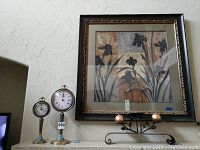 Photo showing large framed botanical wall art with two clocks and candle holder underneath.