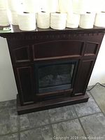 Full view of the electric fireplace shaped like a traditional wood mantel fireplace with an electric insert. Shows dark brown wood with panel details.