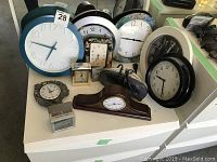 Photo showing assortment of various clocks placed on a white surface including mantel, wall, and alarm clocks