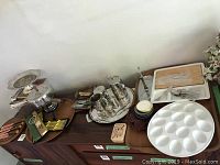 Overview of assorted service ware including silver-plated trays, tea set, egg plate, cutlery, and kitchen utensils arranged on furniture.