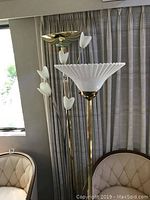 Three floor lamps standing together in front of curtains; two brass lamps with white tulip-style glass shades, one silver floral shade lamp in the middle.