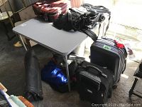 Overview of folding card table with various luggage on and around it under sunlight; includes two rolling suitcases, several duffle bags in black and blue.