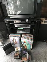 Sony CRT TV on black TV stand with glass doors, vinyl records and CDs in front, two black floor speakers to the side.