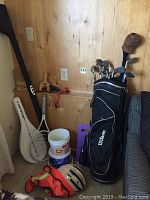 Overall view showing Wilson golf bag containing golf clubs, Dunlop tennis racket and case leaning against the wall, fishing helmet and balls on floor.