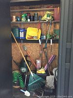 Full view of gardening tools including rake, shovel, hand trowels, clippers, watering can, hose, pots, and bags of topsoil arranged on shelves and floor.