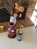 Four oriental style vases grouped on a beige surface including a red Menko vase on wooden base, a cloisonné vase, a large blue and white porcelain vase filled with dried flowers, and a small ginger jar.