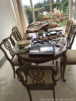 Oblong dining table topped with various linens and accessories, eight chairs around it, sunlit room and garden visible through windows.