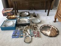 Overview of all silver plated tableware items arranged on a carpet including platters, napkin rings, spoons, bowls, coasters, bread tray and fruit bowl with utensils.