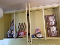 Two irons, handheld steamer, Tide Buzz stain remover, wooden accordion wall hooks, floral ironing board, pillows and drapes stored on a shelf.