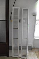 Two tall narrow white framed side lite windows leaning against a wall indoors. Marks visible on frames.