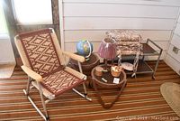 Photo showing vintage rocking chair with patterned seat and back, small tables, globe, lamp, basket of shells, and other small furniture arranged on striped carpet in enclosed patio space.