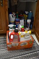 Cleaning products in box and on carpet in front of cabinet showing various bottles including spray bottles and cleaning liquids