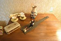 Photo of two Jamaican cups and saucers, silver-plate decorative box, ceramic vase with floral design, wooden dancing marionette on string platform, cheese knife
