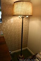 Full view of tall metal floor lamp with beige drum shade turned on, placed in a corner of the room.