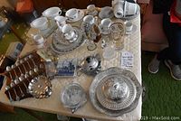 Full view of table with collector spoons on wooden rack, various china cups and saucers, glassware and serving trays on quilted tablecloth.