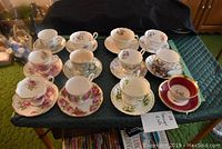 Photo showing all twelve tea cup and saucer sets arranged on a dark green quilted surface, displaying variety of floral patterns and colors, some with gold rims.