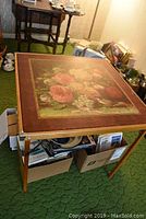 Full view of vintage folding wooden card table with floral design on top, displaying metal hinges and box items beneath.