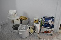 Photo showing full assortment of appliances including boxed Black & Decker chopper, boxed Proctor Silex mixer, Panasonic iron, coffee maker, slow cooker, kettle, and toaster grouped together on floor.