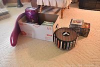 Overall lot including purple Simply Fit Board, box with DVDs, stacks of CDs, and wooden CD carousel on carpet.