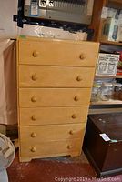 Front view of wooden highboy dresser with six drawers in light wood finish, located in a cluttered space with miscellaneous items around.