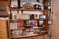 Wide view of five shelves holding assorted kitchen items such as jars, tins, pots, plastic containers, and appliances.