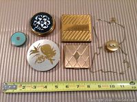 Full view of all five vintage compacts and the eyeglasses chain with container laid on striped fabric with measuring tape for scale.