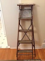 Full front view of wooden step ladder showing four steps, metal braces, worn wood with paint spots, standing against a wall on wooden floor.