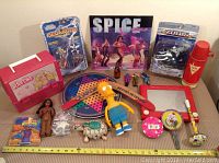 Full view of assorted vintage toys including Barbie lunchbox, Medabots figures, Spice Girls record, Simpsons figure, board game, and more arranged on a table.
