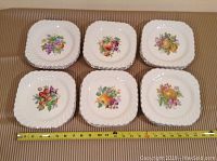 Overview of 24 square white ceramic plates with scalloped edges and various fruit designs clustered together with measuring tape showing approximately 8.5 inches diameter each plate.