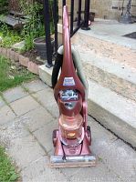 Full view of the upright vacuum cleaner, showing its reddish-pink color, hose, and wheels.