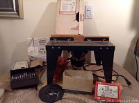 Photo of the Craftsman router table showing metal legs, wooden platform, user manuals, electrical cord, and boxes labeled for bits.
