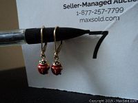 Pair of 14k gold leverback earrings hanging on a pen, showing the ladybug pendant design with red enamel and black spots.