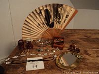 Large wooden hand painted fan, two small red resin Buddha figurines, small white miniature cup, small glass bowl, wooden trinket box, miniature penny bike, round mirror with butterfly detail, necklace, black hair band, heart-shaped glass dish with broken jewelry, earrings, miniature keys with lock all arranged on wood table.