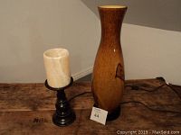 Two table lamps on wooden table, showing a tall wood grain style lamp and a smaller lamp with black base and off-white cylindrical shade. Lamps are unplugged.