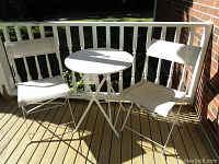 Photo of white metal round folding table and two white metal folding chairs with waterproof fabric seats and backs on wood deck.