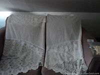 Two Jardiniere lace curtains displayed on a sofa, showing detailed lace floral pattern and scalloped edges.