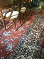 Photo showing area rug under dining table and chairs, revealing reddish floral pattern and detailed cream border.