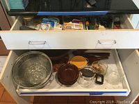 Two open drawers showing assorted kitchen bowls in wood, glass, and metal strainers, plus wood utensils.