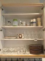 Wide view of white cabinet shelves containing plates stacked, various clear stemmed and cordial glasses, white paper cups, ceramic tea pot, ramekins, and a small green and white bud vase.