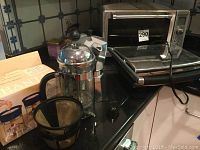 Photo shows a Krups panini press, French press, moka coffee maker, plastic measuring cups, and a coffee filter on a kitchen countertop along with boxes.