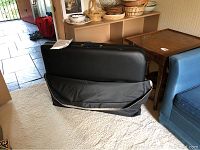 Portable massage table folded inside black carry bag, placed on carpet floor with some furniture in the background.