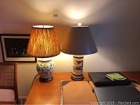 Two ceramic table lamps placed on a wooden desk, both turned on to show illumination. One lamp has a round floral decorated base and the other a cylindrical Asian-themed design base.