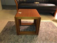 Three teak nesting tables stacked inside each other on floor in front of leather sofa, showing compact nest and finish.