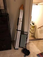 Photo of folded 4 foot ladder leaning against wall near dark furniture with folding stool and knee pads on floor.
