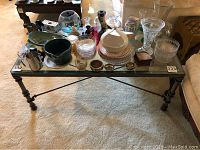 Rectangular coffee table with glass top and metal legs supporting an assortment of glassware, ceramics, and decorative household items, placed on a beige carpet.