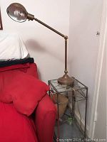 Photo of vintage brass tensor lamp sitting on top of iron framed glass table next to red upholstered furniture.