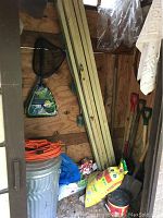 Interior of garden shed showing 2x4 lumber, shovels, bags of garden chemicals, trashcans, extension cord, and pond nets.
