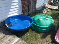 Two items outdoors on grass and deck: a green frog-shaped covered sandbox and a blue round kiddie pool.