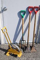 Roofing tools shown outside against a white door, including two red-handled shingle removers, one green-handled roofers pitch fork, and a yellow metal hoisting wheel.